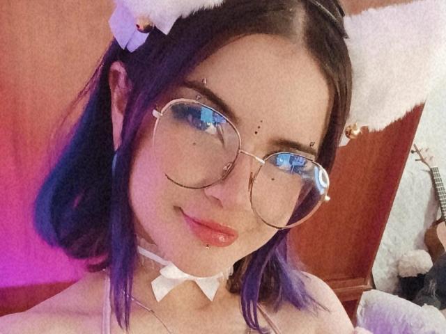 RavenThuwu with purple hair, cat ears, round glasses, nose ring, and a white bow choker, smiling at the camera, showing red lips and a slight blush.