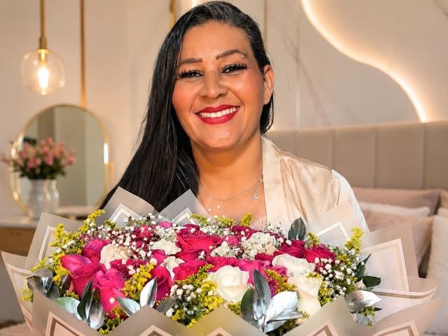 VictoriaKiley with long black hair, smiling, wearing red lipstick, holding a bouquet of pink, white, and yellow flowers with green leaves. She's wearing a white shirt.