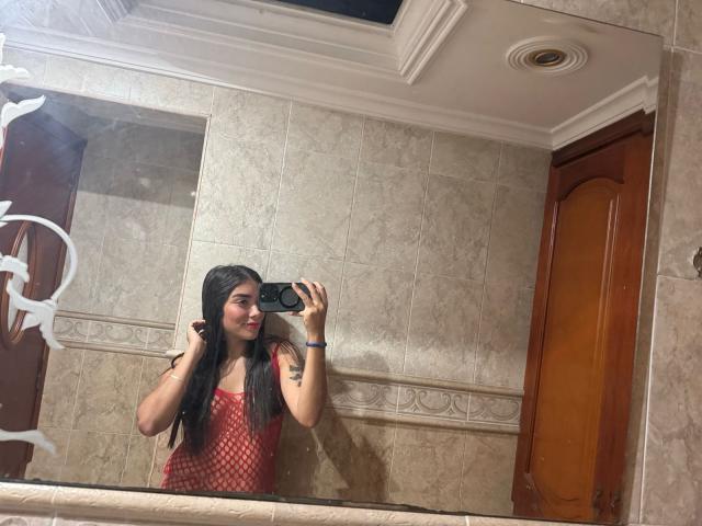 SharonKlart with long black hair, red fishnet top, holding phone, smiling, tattoo on right arm, brown tiled bathroom, mirror reflection, white ceiling, wooden door, blue wristband.