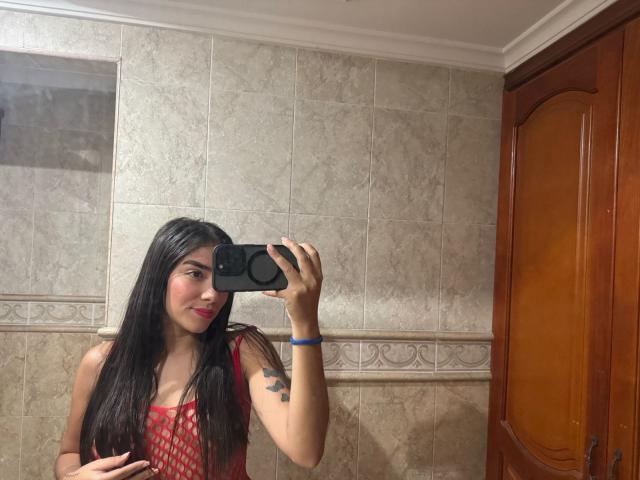 SharonKlart with long black hair, red dress, pink lipstick, holding phone, taking mirror selfie, visible tattoo on left arm, wooden cabinet on right, blue bracelet on left wrist.