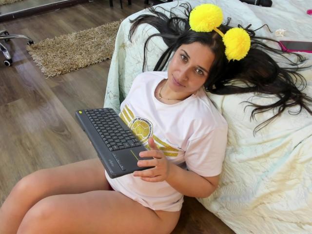 EmmyRossi with yellow pom-pom cat ears, sitting on wooden floor, wearing white tee and no pants, holding black laptop, long dark hair, small ass, smiling, bedroom.