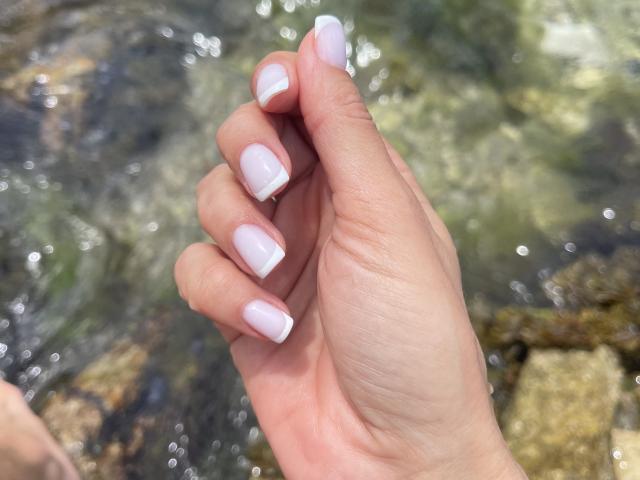 SandraSwweet's hand with light pink French-manicured nails, showing off her clean, smooth, pale skin against a blurry water background.