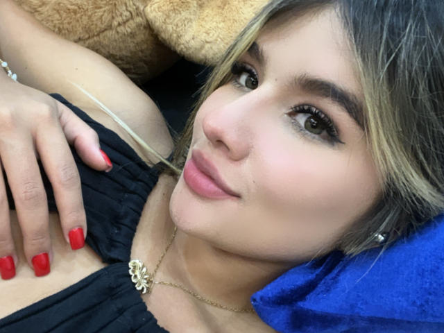 SashaPolet with blonde hair, red nails, and black top, lying down, smiling, showing small tits, gold necklace, blue pillow, and brown teddy bear in the background.