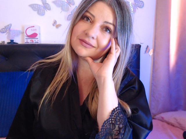 NatalyHami with blonde hair, black lace sleeves, and a slight smile, resting her hand on her cheek, wearing a black satin robe, in a softly lit room.