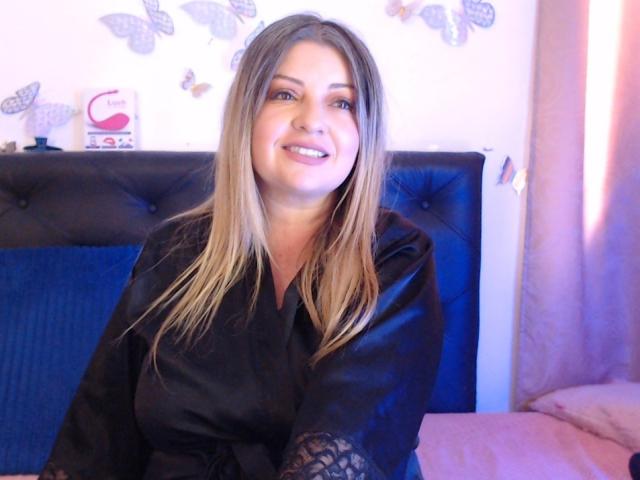 NatalyHami with blonde hair, smiling, wearing a black satin robe with lace sleeves, sitting on a blue tufted headboard. Boobs partially visible under the robe.