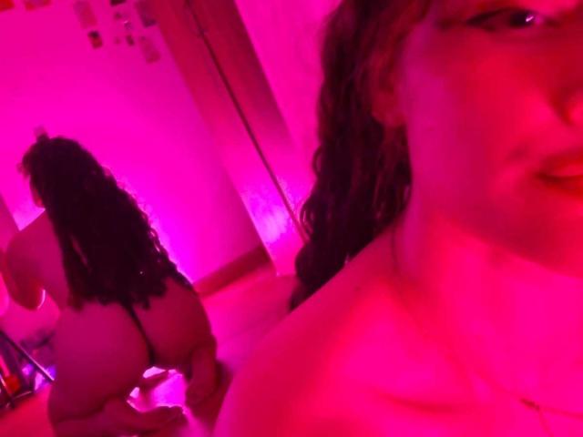 DanaStone with curly black hair, nude, butt facing camera, pink-lit room, DanaStone's face in foreground, slightly blurred. DanaStone's ass, back, and reflection visible.