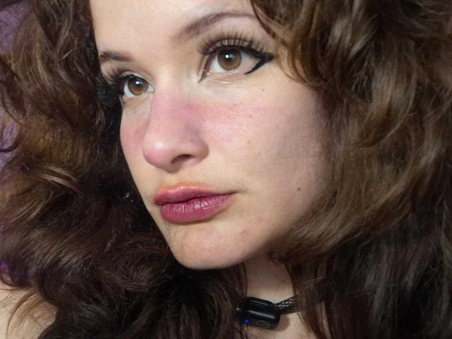 DanaStone's face, showing curly brown hair, brown eyes with black eyeliner, pink lips, slightly flushed skin, and wearing a black choker with a blue bead.
