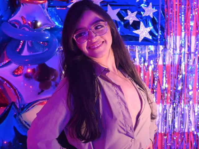 EvaMcAllister with long black hair, glasses, smiling, wearing an open grey shirt, showing her tits, standing in a colorful, neon-lit, party setting with shiny blue and silver decorations.