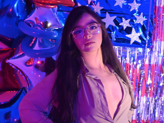 EvaMcAllister with long dark hair, wearing glasses, unbuttoned shirt revealing her tits, standing against colorful, festive background with balloons and stars, looking directly at the camera.