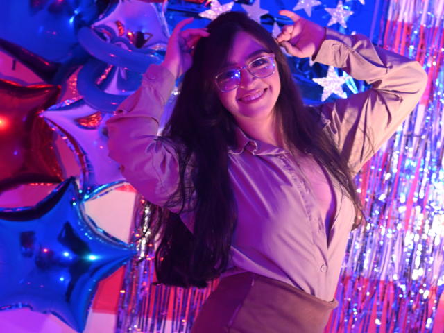 EvaMcAllister with long black hair, glasses, smiling, hands in hair, wearing beige shirt, brown pants, standing against colorful star and ribbon background, blue and purple lights.