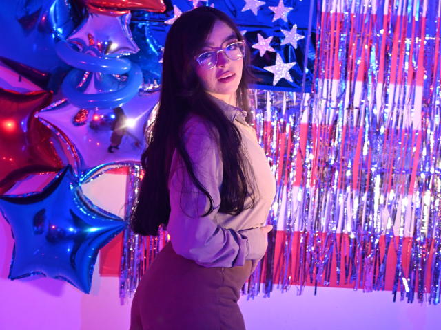 EvaMcAllister with long black hair, glasses, and a cute smile, wearing a beige top and brown pants, standing in front of colorful American flags and shiny streamers.