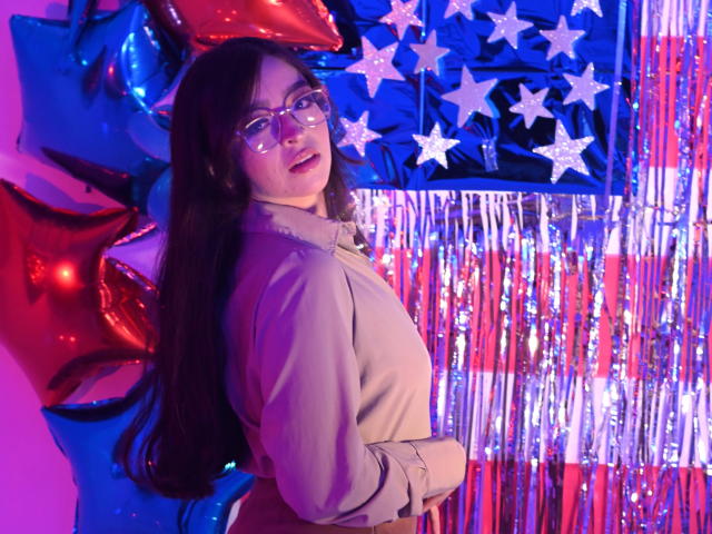 EvaMcAllister with long dark hair, glasses, and a beige jacket, standing with her back slightly turned, looking over her shoulder. Red and blue balloons, star-shaped decorations, and silver streamers.