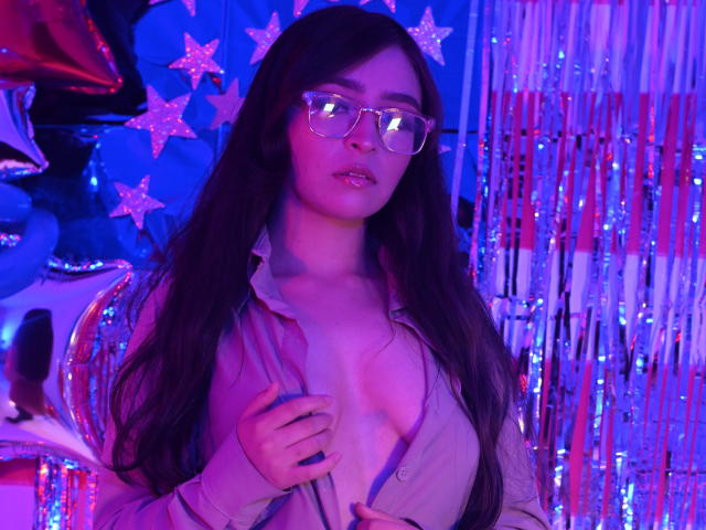 EvaMcAllister with long dark hair, glasses, and pink shirt open to reveal small tits, standing in purple and blue neon light, holding shirt. Background with star and strip decorations.