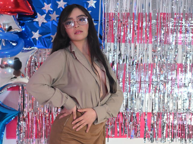 EvaMcAllister with long black hair, glasses, beige blouse exposing her big tits, brown pants, hand on hip, standing in front of American flag-themed decor with silver and red streamers.