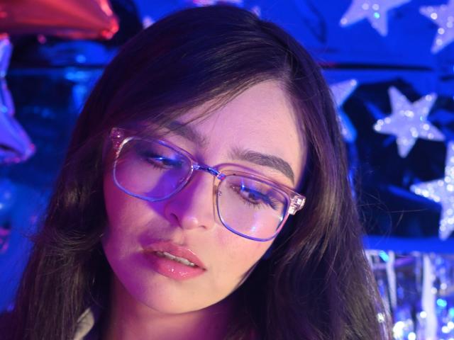 EvaMcAllister with dark brown hair, purple glasses, and pink glossy lips, looking down with a slight pout, blue and purple light reflecting on her face.