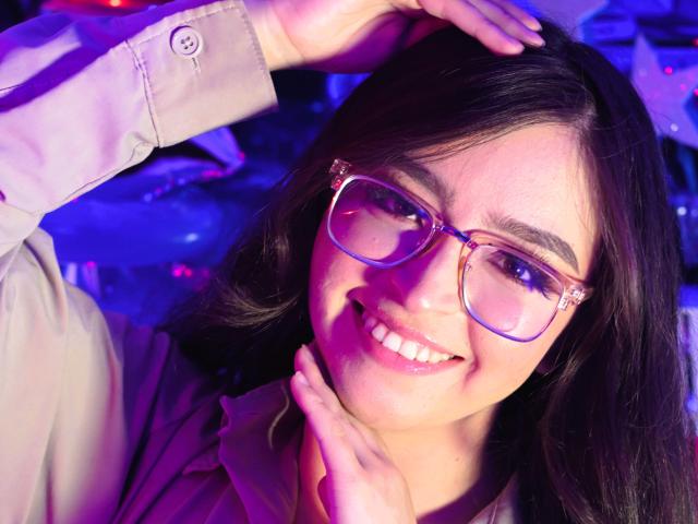 EvaMcAllister with glasses, smiling, hands on head and neck, wearing a beige shirt, dark hair, blue-purple light, clear skin, cute, sexy, relaxed, happy expression.