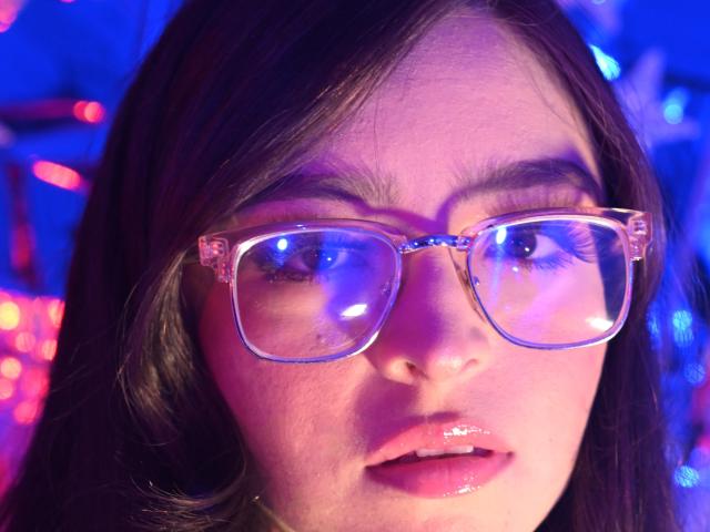 EvaMcAllister with shiny pink lips and reflective clear glasses, blue and purple lights reflecting in her eyes, brown wavy hair, slightly parted pink glossy lips.