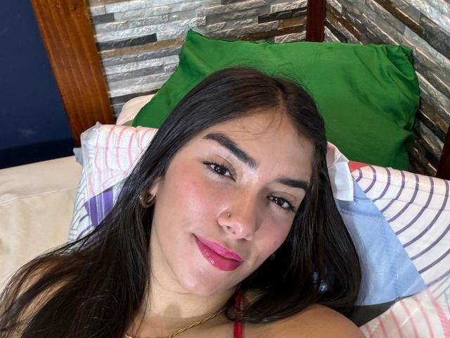 SharonKlart has long black hair, brown eyes, pink lipstick, and a slight smile. She's lying on a bed with green and striped pillows, wearing a red top.