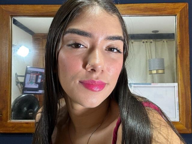 SharonKlart has long brown hair, a nose ring, and pink lipstick, smiling at the camera with her tits barely visible in a red top, reflected in a wooden mirror.