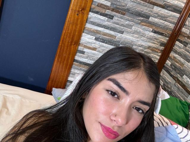 SharonKlart with long black hair, pink lipstick, and a nose ring, smiling slightly, looking at the camera, showing her ass in the background, on a bed with pillows.