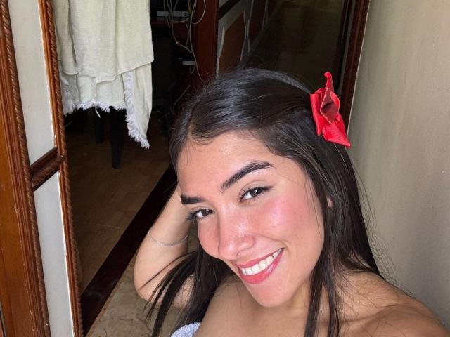 SharonKlart with dark brown hair, red hair clip, smiling, pink lips, bare shoulders, visible tan skin, and brown eyes, holding her hair. Boobs not visible.
