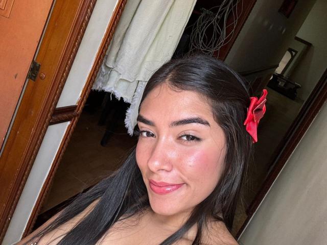 SharonKlart with shiny pink lips, black straight hair, and a red hair clip, smiling at the camera, showing off her cute, slightly flushed face.