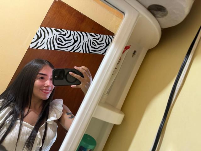 SharonKlart with long black hair, smiling, taking a selfie in a mirror, holding a black phone, wearing a white ruffled shirt, showing a tattoo on her left arm.