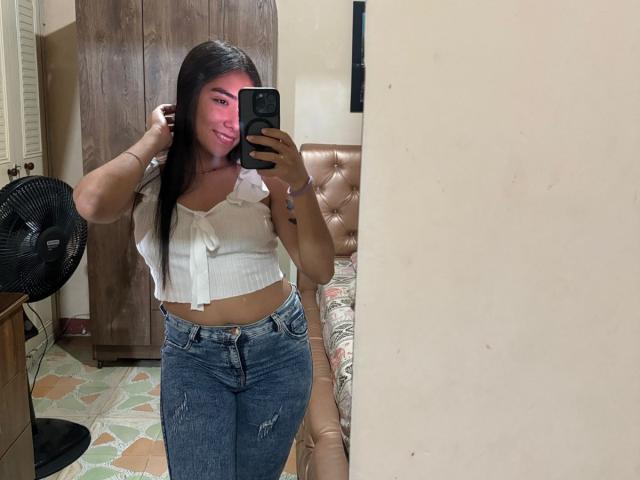 SharonKlart with long black hair, smiling, wearing a white ribbed crop top and blue ripped jeans, taking a mirror selfie with a pink face filter.