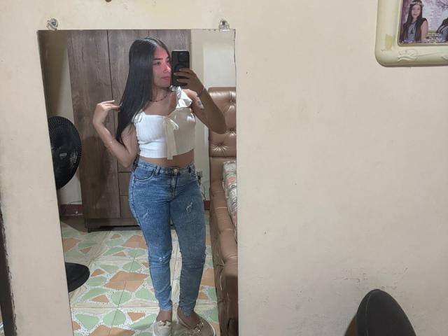 SharonKlart with long black hair, white strapless crop top, and blue jeans, standing in front of a mirror, holding her hair, showing her small tits and slim ass.