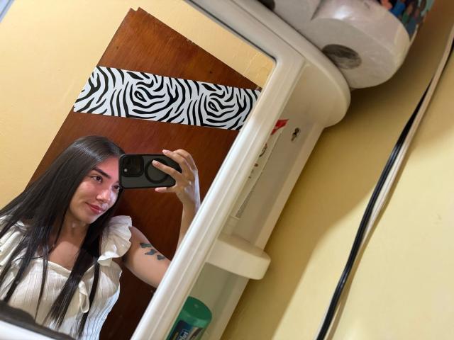 SharonKlart with long black hair, small tits, white blouse, holding phone, mirror selfie, zebra-striped door, tattoo on arm, yellow wall, toilet paper roll, smiling, cute.