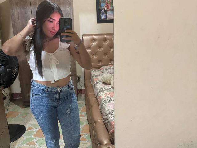 SharonKlart with long dark hair, pink face filter, white crop top, blue ripped jeans, holding phone, mirror selfie, tan tufted bed, black fan, beige wall, and patterned floor.