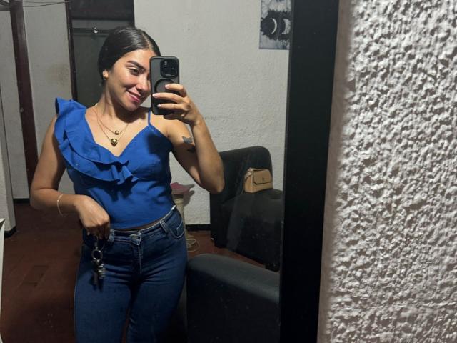 SharonKlart with medium tits, blue ruffle top, dark jeans, holding phone in mirror selfie, smiling, black hair, necklace, keys in right hand, white textured wall in background.