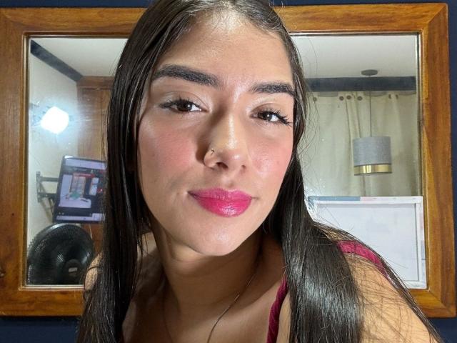 SharonKlart with long black hair, pink lipstick, and brown eyes, wearing a red top, smiling slightly, reflection in wooden mirror, medium skin tone, subtle makeup, black eyelashes.