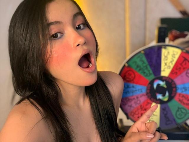 MiiaPalmer with long black hair, wide eyes, and open mouth, showing her pink tongue, pointing at a colorful spin wheel, topless, with visible tits.