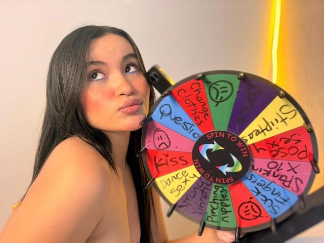 MiiaPalmer with long black hair, pouty lips, and a bare shoulder, holding a colorful spin-the-bottle game with "SintToDirty" logo, looking to the side.