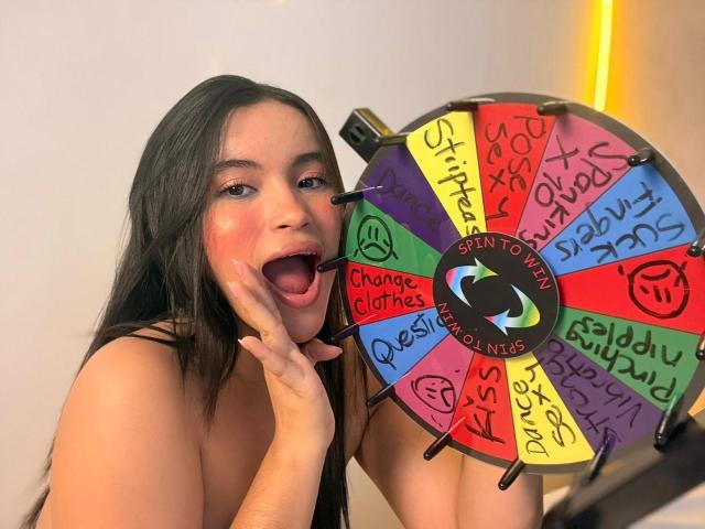 MiiaPalmer with long dark hair, blush on cheeks, open mouth, holding a colorful spin wheel, touching her chin, wearing nothing, looking playful and sexy.