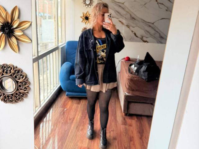 ElenaSaenz with blonde hair, black jacket, graphic tee, beige skirt, black tights, and black boots, taking a mirror selfie, showing her ass and tits slightly visible under the skirt.