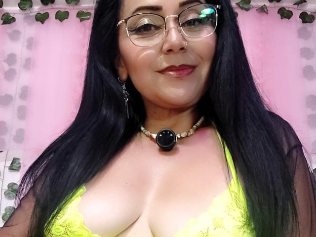 Zaria69 with long black hair, glasses, and nose ring, wearing a yellow lace bra, necklace with black gem, and sheer top, smiling at the camera, showing her tits.