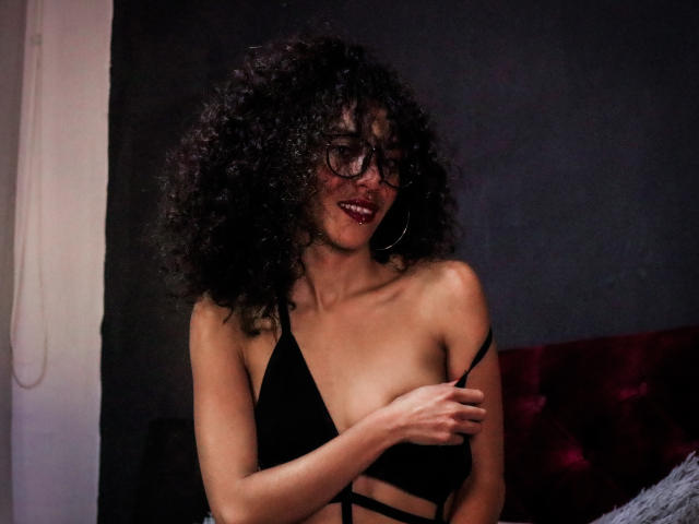 LiliMuller with curly black hair, glasses, and red lipstick, wearing a black strappy top, slightly covering her tits with her hand, smiling seductively.