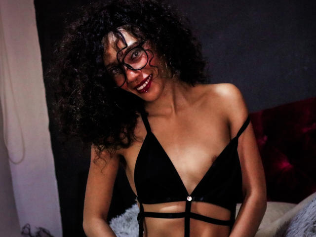 LiliMuller with curly black hair, wearing black strappy bra, smiling, red lipstick, glasses, showing small tits, dark skin, standing in dimly lit room.
