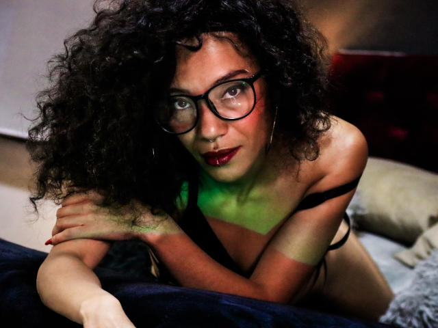 LiliMuller with curly black hair, glasses, red lipstick, and black strappy lingerie, leaning on a bed, green light on her chest, looking at the camera with a sexy gaze.