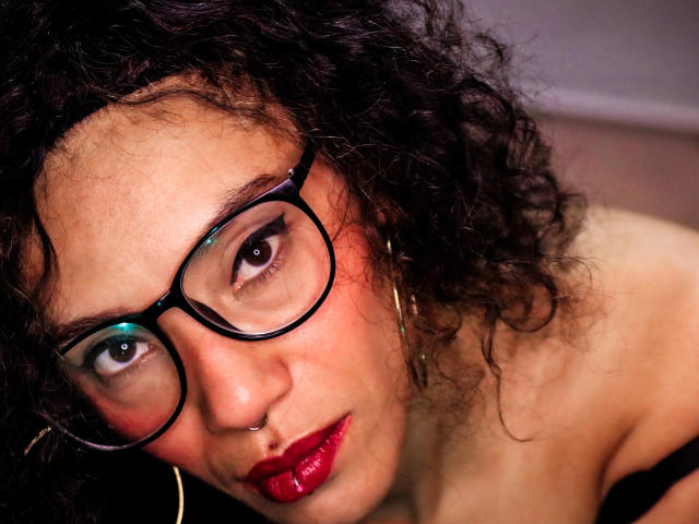 LiliMuller has curly dark hair, glasses, red lipstick, nose ring, and hoop earrings, looking up with a slightly serious expression. Her tits are not visible.