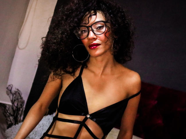 LiliMuller with curly black hair, red lipstick, glasses, large hoop earrings, and a black strappy bra, showing her tits and toned abs, looking sexy and confident.