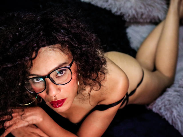 LiliMuller with curly black hair, glasses, red lipstick, black bra, and thong, lying on a furry black and gray surface, looking up with a serious expression, showing her tits.