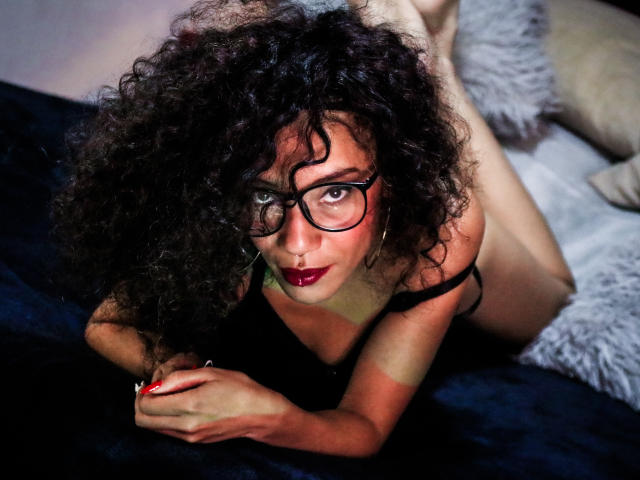 LiliMuller with curly black hair, red lipstick, glasses, and black lingerie, lying on a dark blue bed, staring seductively at the camera, legs bent behind her.