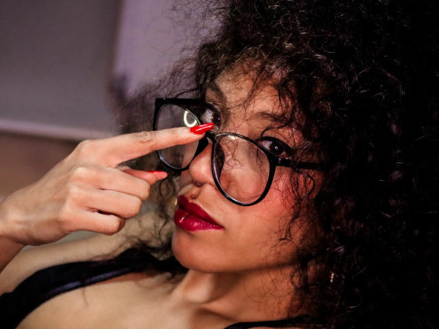 LiliMuller with curly black hair, wearing black glasses, red lipstick, and red nails, touching her glasses with her index finger, looking slightly to the side.