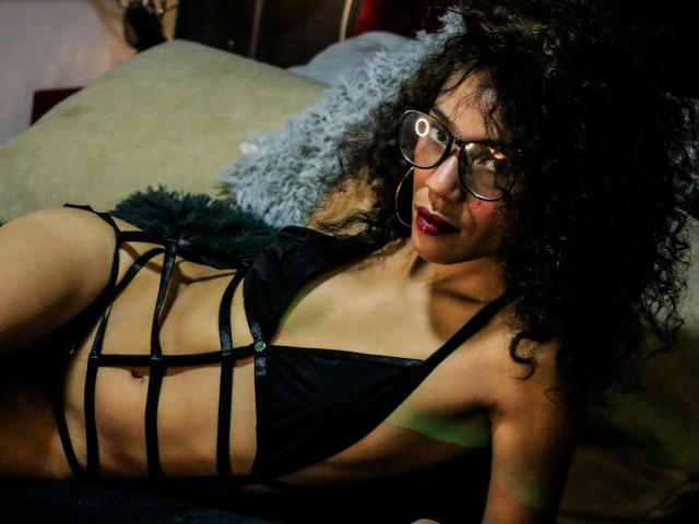 LiliMuller with curly black hair, wearing glasses, black strappy lingerie, showing her tits and flat stomach, lying on a bed, looking at the camera with red lipstick.