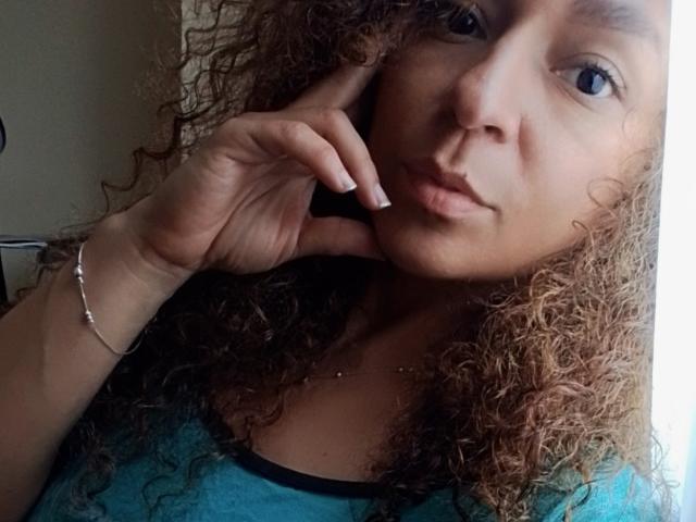 Angelonix with curly brown hair, blue eyes, and tan skin, touching her cheek, wearing a teal shirt, and a silver bracelet, looking slightly pouty. Boobs visible under shirt.