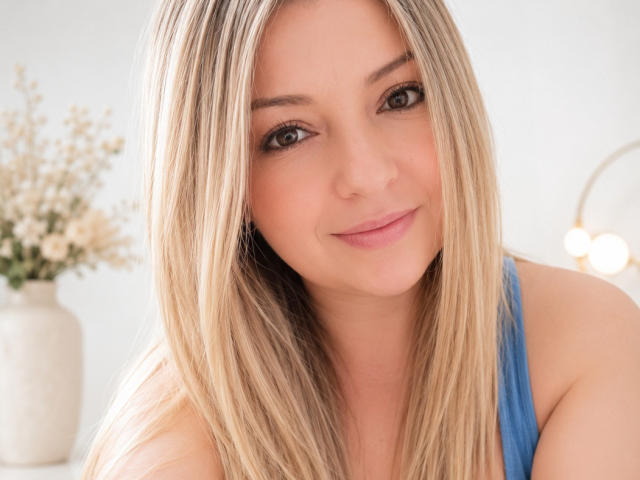 NatalyHami has blonde hair, blue eyes, and a slight smile. She's wearing a blue top, showing her bare shoulders. Her tits are small and her skin is fair.
