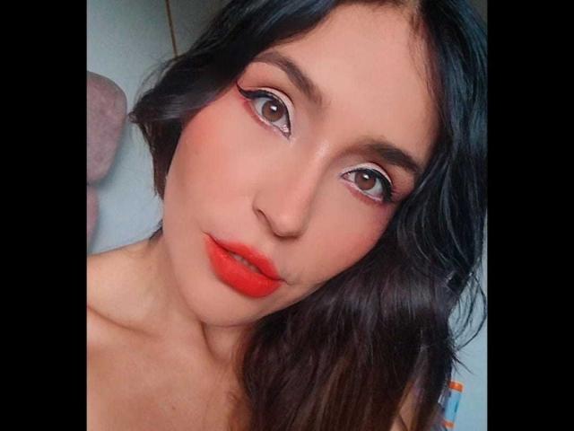 MindyLove's face, with red lipstick, black wavy hair, and bold eyeliner, showing her brown eyes and slightly parted red lips in a selfie. Her tits are not visible.